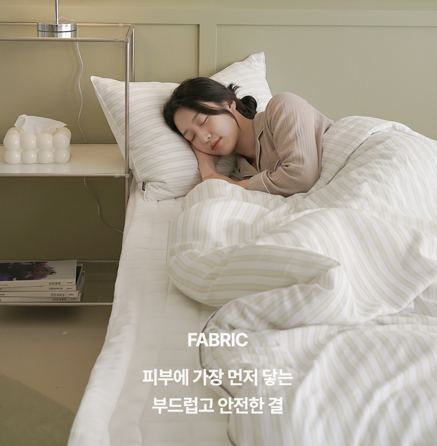 FABRIC 피부에 가장 먼저 닿는 부드럽고 안전한 결 - 옐로우 그린 스트라이프 패턴의 누비지오 모스트(MOST) 차렵이불을 덮고 수면을 취하는 여성 모델 연출컷, 원단 감촉 강조 메인 섹션